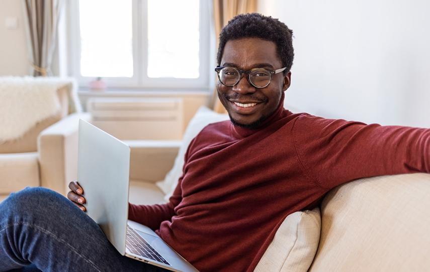 Black man in red shirt and glasses smiles while sitting on couch with a laptop