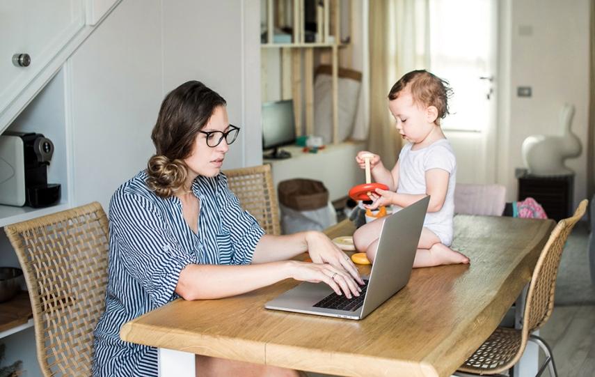 Woman works on laptop on table with toddler next to her